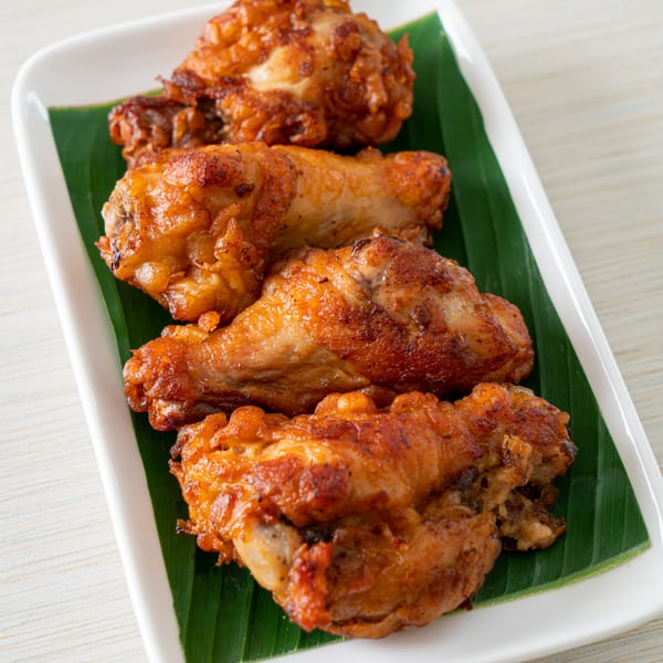 Four crispy, golden-brown fried chicken pieces on a white plate with a green banana leaf.